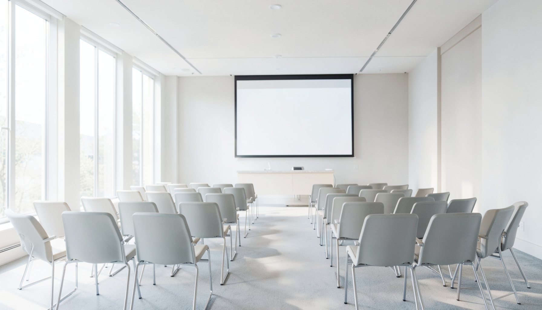 Sala de conferencias luminosa con sillas dispuestas en filas, una pantalla de proyección al fondo y luz natural entrando por amplios ventanales, en tonos neutros y cálidos.